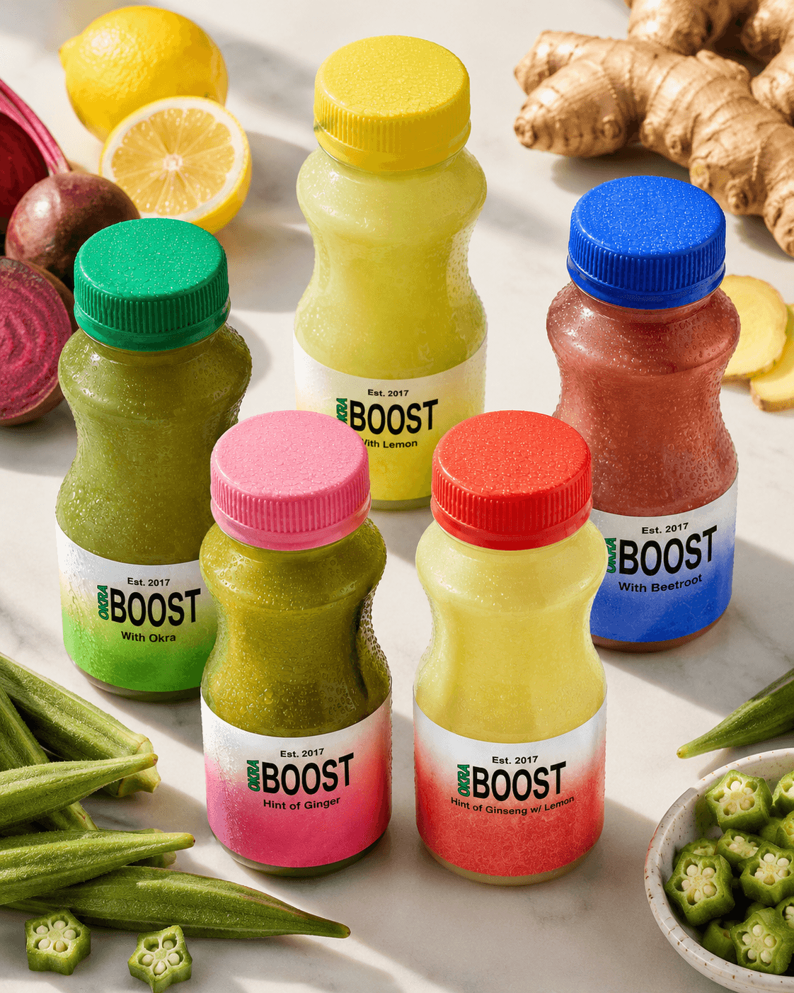 All five Okra Boost flavors on a marble counter alongside their raw ingredients — whole okra pods, sliced lemon, ginger root, beetroot, ginseng
