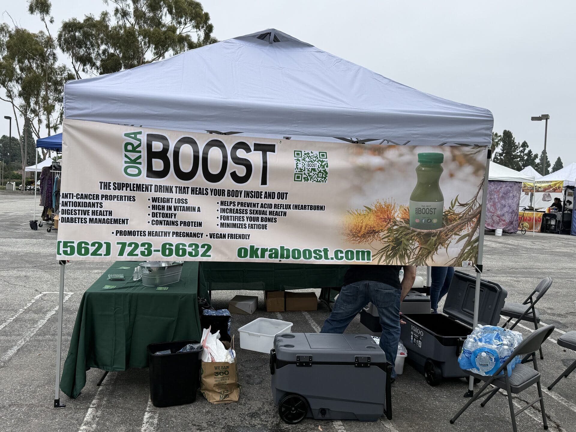 A wide shot of the OKRA BOOST farmers market booth — the bilingual banner above the table reads Anti-Cancer Properties, Antioxidants, Digestive Health, Diabetes Management, with the bottle and phone number visible in front