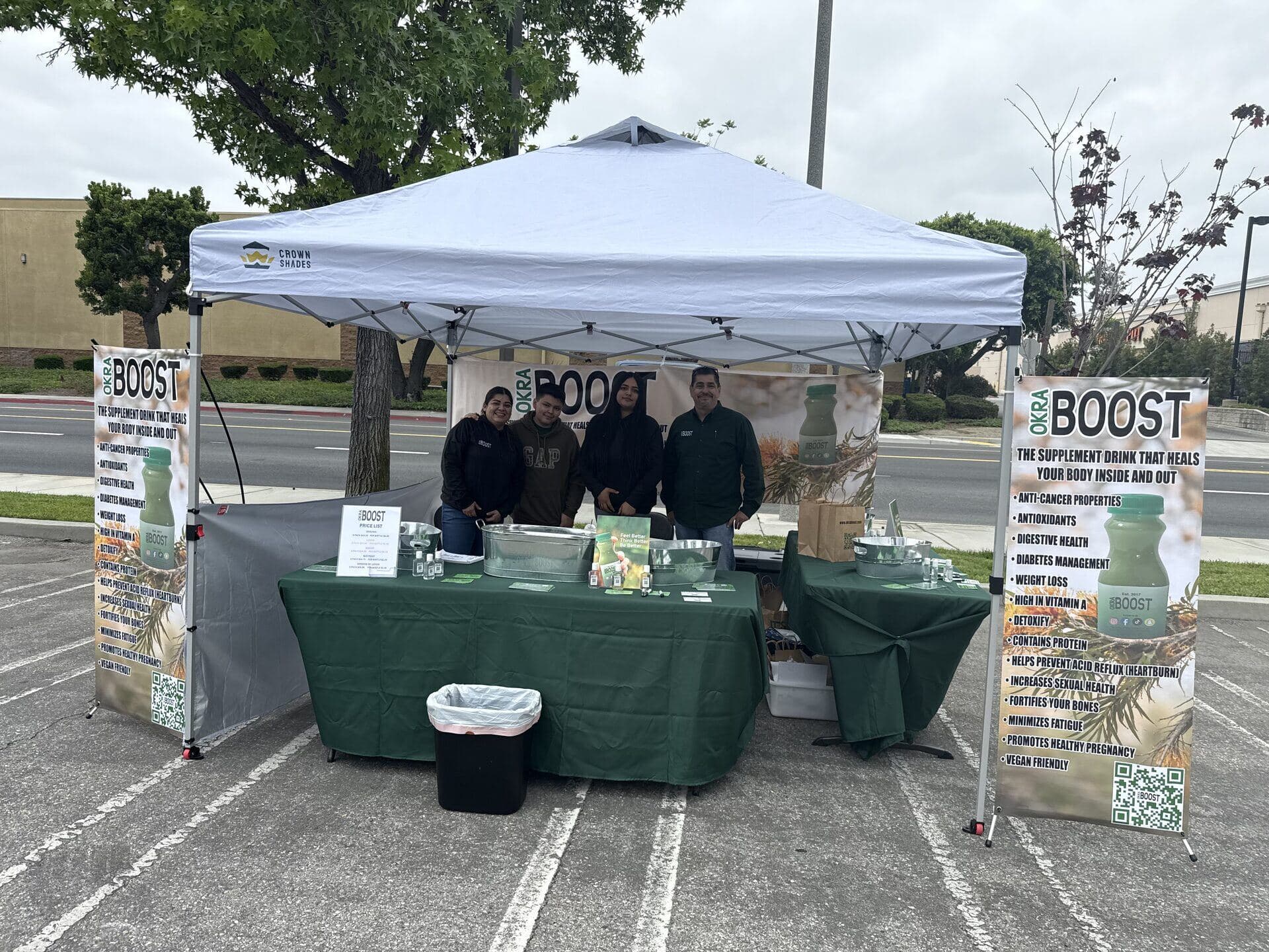 The OKRA BOOST farmers market booth — white pop-up tent in a Long Beach parking lot, four team members standing under it next to coolers of bottles and a tall banner of product benefits