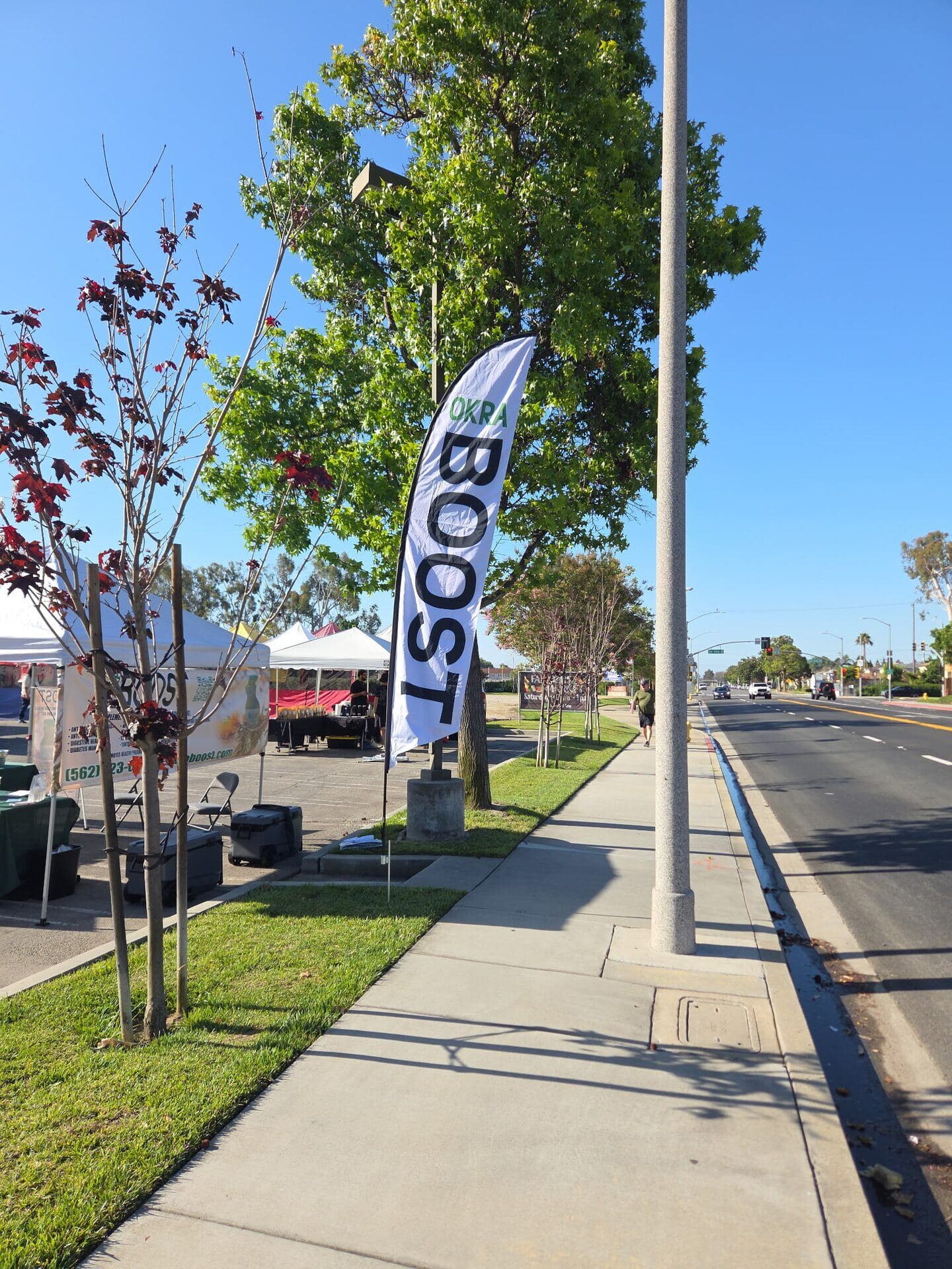 The OKRA BOOST tall sail-banner planted on the sidewalk along a Long Beach street — the white tent and tables of the booth visible behind it under a bright blue sky
