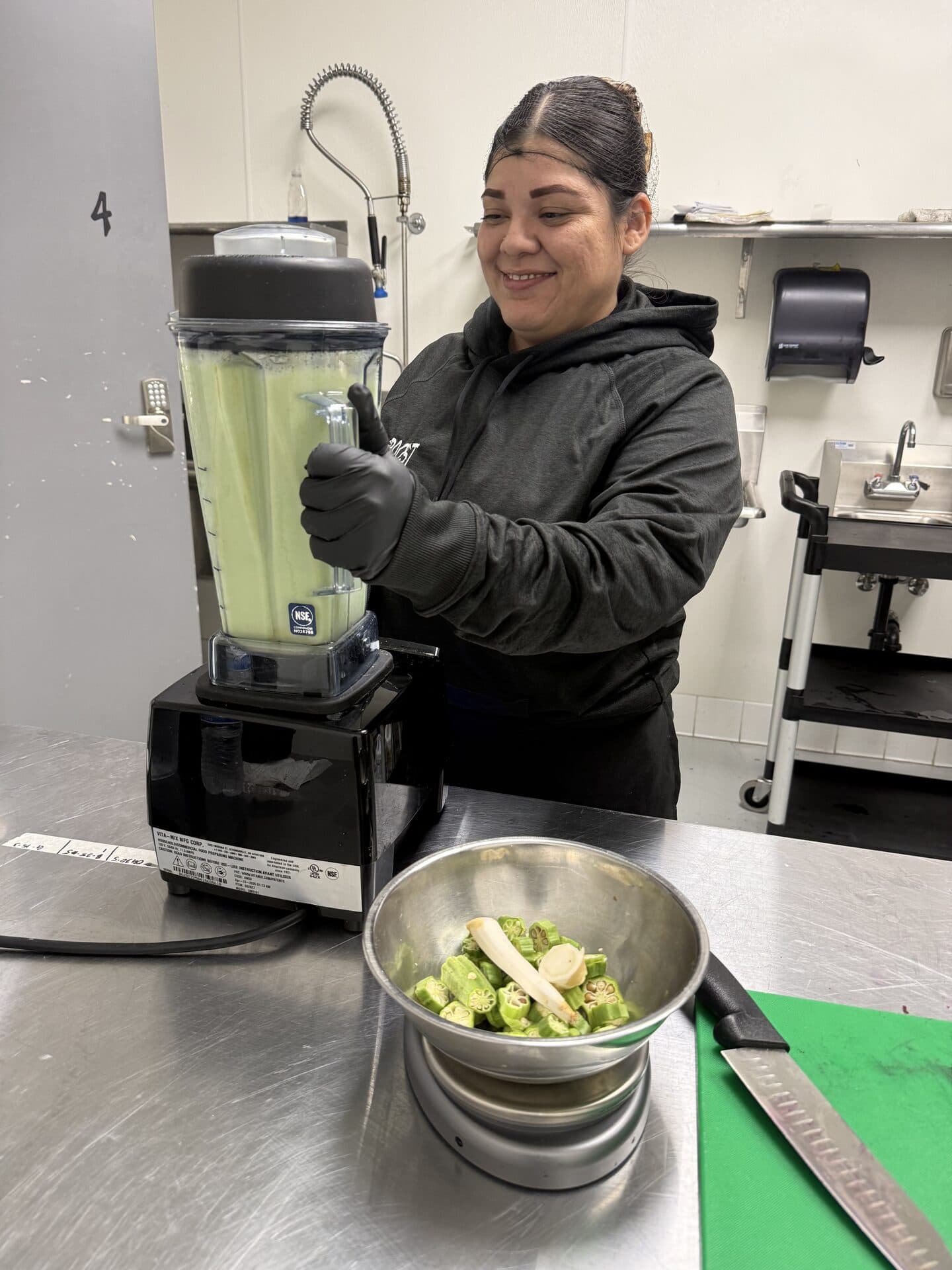 A team member in an Okra Boost hoodie blending a fresh batch of green okra in the Long Beach kitchen, with a bowl of sliced okra and ginger waiting on the stainless prep table