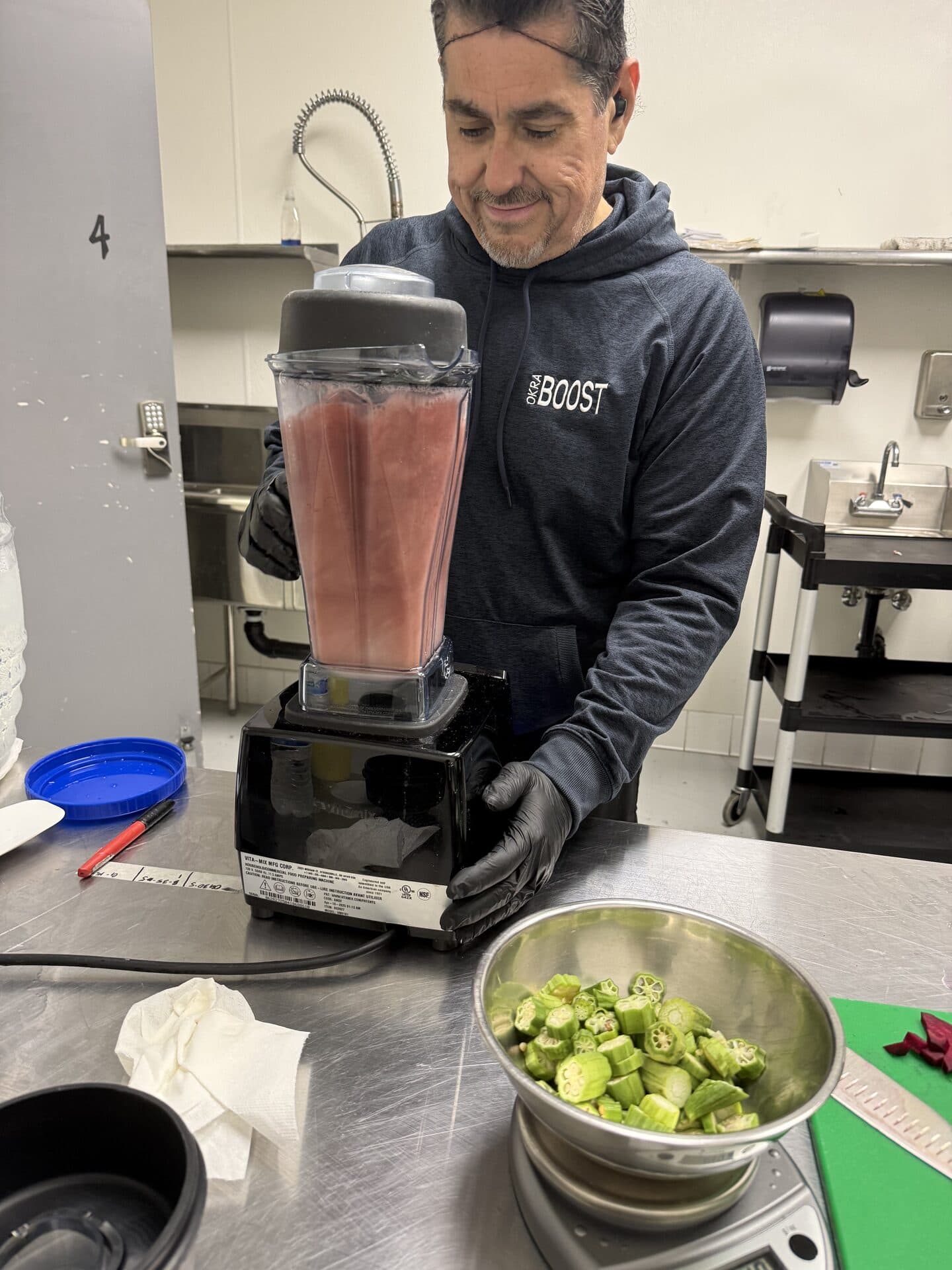 A member of the OkraBoost team in a branded hoodie running the Beetroot blend through the kitchen blender, with a fresh bowl of sliced okra on the prep table beside him