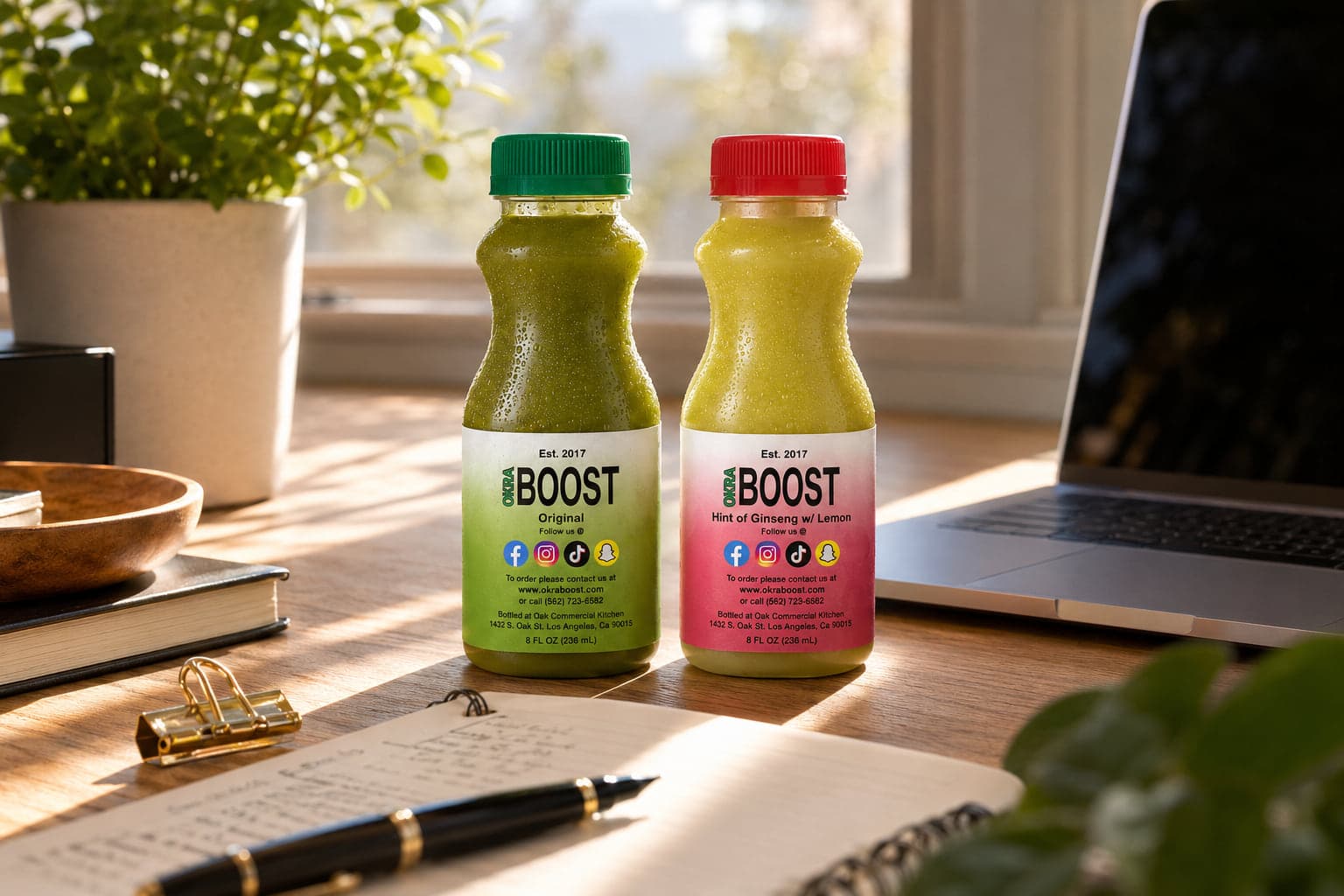 Two Okra Boost bottles — Original and Ginseng-Lemon — on a sunlit desk beside an open notebook, a laptop, and a small house plant